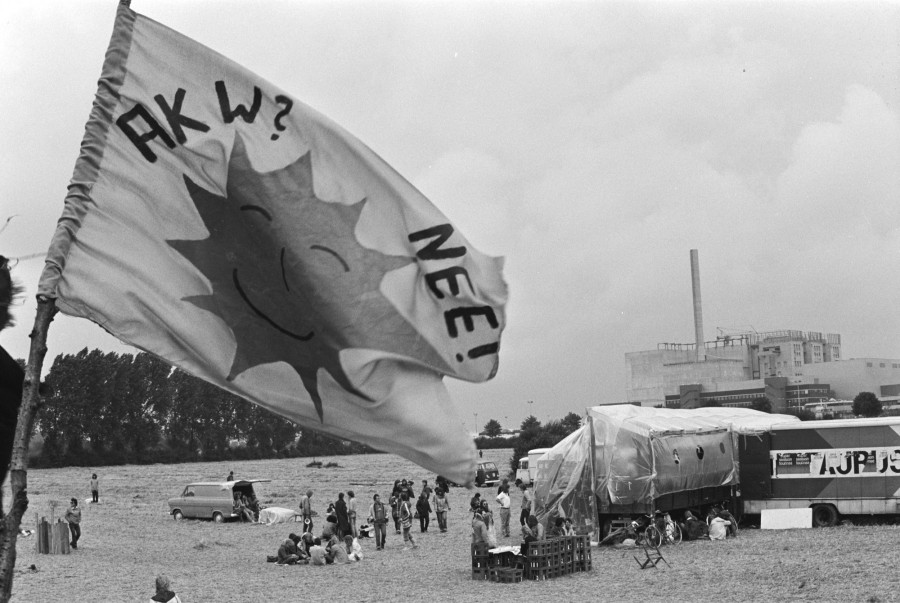Im Vordergrund weht eine Fahne, auf der eine lachende Comic-Sonne abgebildetet ist, mit dem Schriftzug "AKW? Nee!". Im Hintergrund das Kernkraftwerk, davor eine Wiese mit vereinzelten Menschengruppen und einer Zeltkonstruktion.