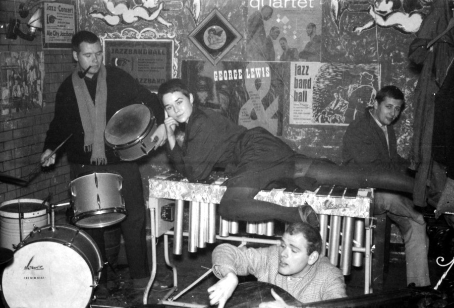 4 Personen mit Musikinstrumenten vor einer plakatierten Wand. Im Vordergrund am unteren Bildrand sieht man einen Mann, der auf einem Kontrabass spielt. Links im Bild ein Mann mit Pfeife im Mund, der stehend eine Snaredrum in der Hand hält. Im Zentrum des Bildes liegt eine Frau mit kurz geschorenen Haaren auf einem Vibraphon. Rechts von ihr hinter dem Vibraphon sitzt ein Mann seitwärts und schaut in die Kamera.